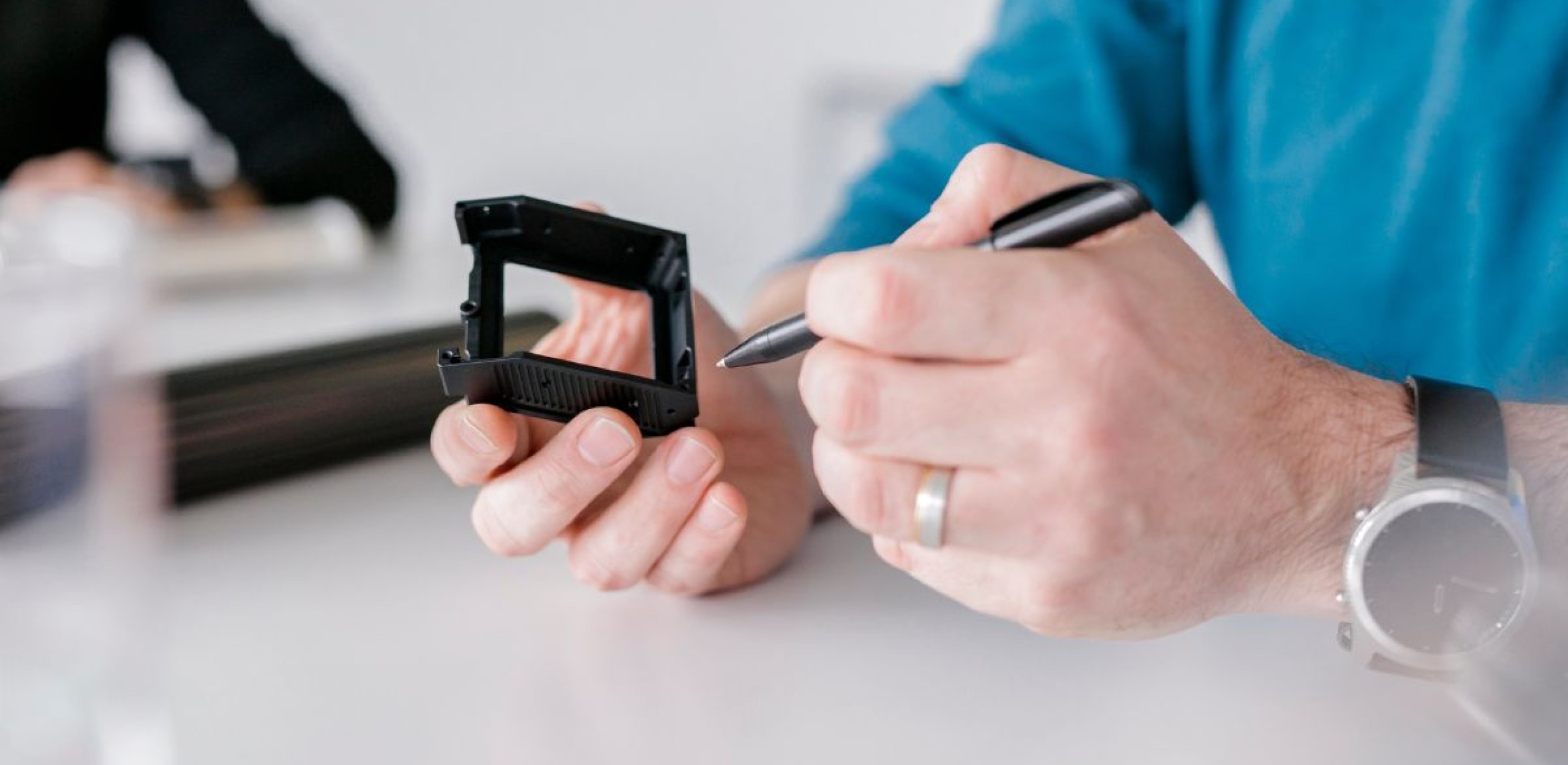 Ein Mann hält ein gefrästest schwarzbeschichtetes CNC Fertigungsbauteil inder Hand und zeigt mit einem Kugelschreiber auf eine Kante des Bauteils im Hntergrund sitzt eine weitere Person und lässt sich von dem Mann beraten