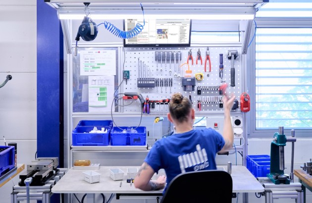 Frau sitzt vor einem Montageplatz und greift nach einem Werkzeug ihr Shirt hat das PWB Logo aufgedruckt und der Arbeitsplatz ist sauber aufgeräumt und gut strukturiert