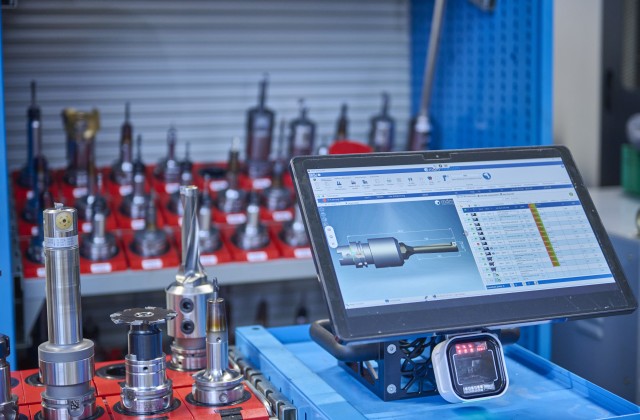 Im Vordergrund ist ein Display zu sehen mit einem Scanner und einem Bild eines Werzeugs mit einer Liste, links daneben und im HIntergrund stehen Werkzeuge für CNC Bearbeitung